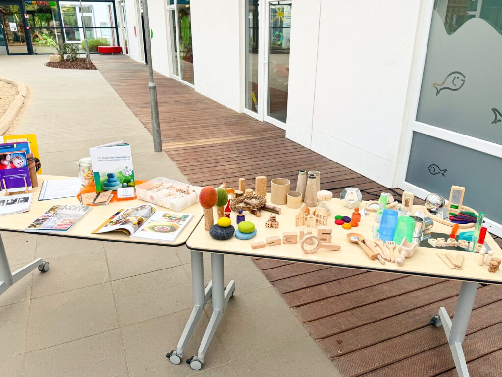 Auf zwei zusammengestellten Tischen in einer Kita sind verschiedene pädagogische Spiel- und Entdeckungsmaterialien aus Natur- und Alltagsmaterialien ansprechend ausgelegt.